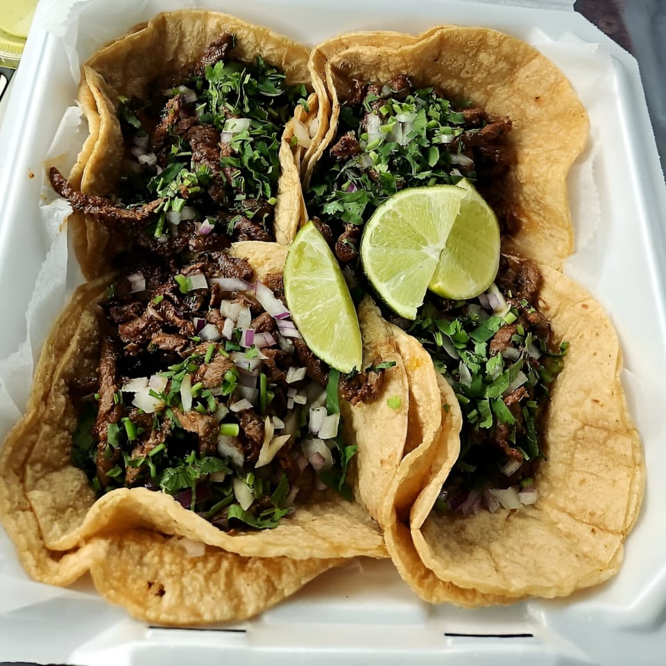 Four Sabor Mexican street tacos with carne asada, cilantro, onions, and lime wedges in a to-go container.